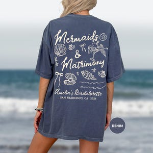 May include: Denim blue t-shirt with white graphic text and illustrations. The text reads "Mermaids & Matrimony" with seashell, bow, and mermaid-themed drawings. Below, it says "Amelia's Bachelorette, San Francisco, CA 2026".