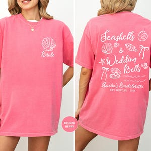 K&ouml;nnte beinhalten: Ein pinkfarbenes T-Shirt mit wei&szlig;em Text und Grafiken. Auf der Vorderseite des Shirts steht "Bride" mit einer Muschel-Grafik. Auf der R&uuml;ckseite des Shirts steht "Seashells & Wedding Bells" mit Muschel- und Schleifen-Grafiken. Der Text auf der R&uuml;ckseite lautet "Amelia's Bachelorette Key West, FL 2026".