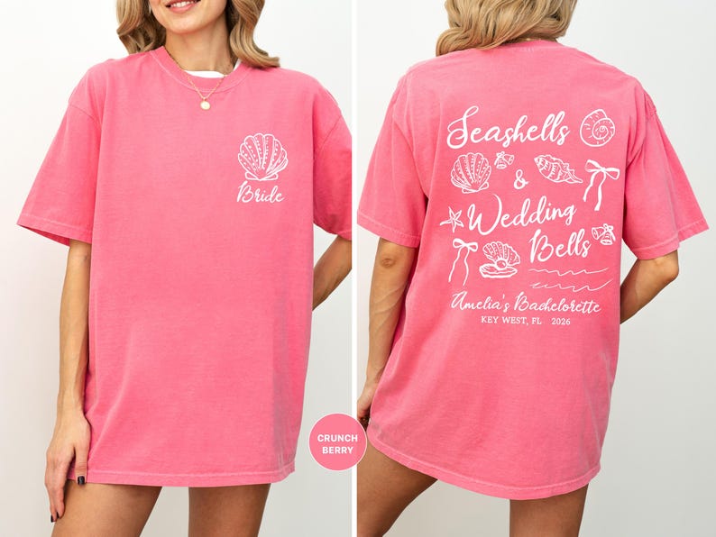 K&ouml;nnte beinhalten: Ein pinkfarbenes T-Shirt mit wei&szlig;em Text und Grafiken. Auf der Vorderseite des Shirts steht "Bride" mit einer Muschel-Grafik. Auf der R&uuml;ckseite des Shirts steht "Seashells & Wedding Bells" mit Muschel- und Schleifen-Grafiken. Der Text auf der R&uuml;ckseite lautet "Amelia's Bachelorette Key West, FL 2026".