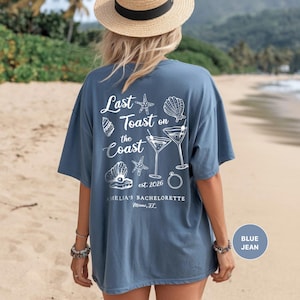 K&ouml;nnte beinhalten: Blaues Jeans-T-Shirt mit wei&szlig;em Text und Grafiken. Auf der R&uuml;ckseite des Shirts steht der Satz "Last Toast on the Coast" mit Muschel- und Cocktail-Illustrationen. Der Text "AMELIA'S BACHELORETTE Miami, FL" ist ebenfalls sichtbar.