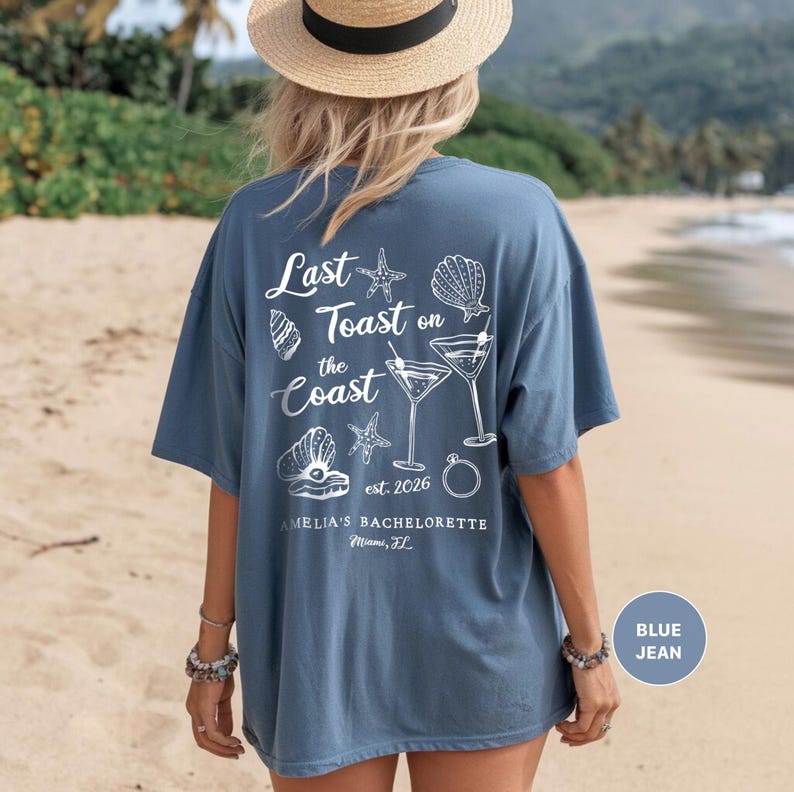 K&ouml;nnte beinhalten: Blaues Jeans-T-Shirt mit wei&szlig;em Text und Grafiken. Auf der R&uuml;ckseite des Shirts steht der Satz "Last Toast on the Coast" mit Muschel- und Cocktail-Illustrationen. Der Text "AMELIA'S BACHELORETTE Miami, FL" ist ebenfalls sichtbar.