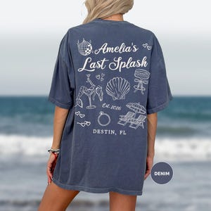 Puede incluir: Una camiseta azul denim con gr&aacute;ficos blancos en la espalda. El texto dice "Amelia's Last Splash" y "Destin, FL". El dise&ntilde;o presenta una bola de discoteca, un c&oacute;ctel, una concha, un anillo y sillas de playa.