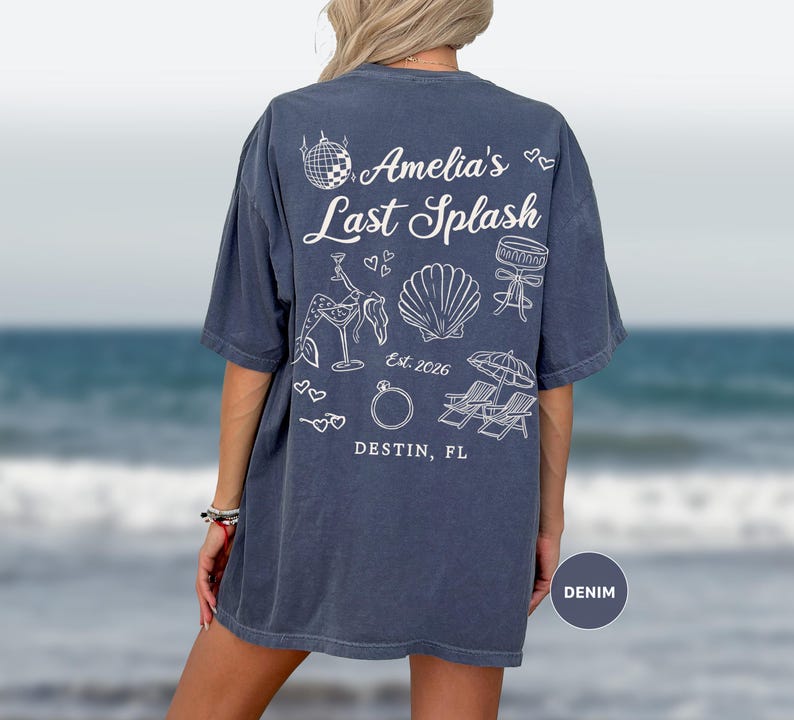 Puede incluir: Una camiseta azul denim con gr&aacute;ficos blancos en la espalda. El texto dice "Amelia's Last Splash" y "Destin, FL". El dise&ntilde;o presenta una bola de discoteca, un c&oacute;ctel, una concha, un anillo y sillas de playa.