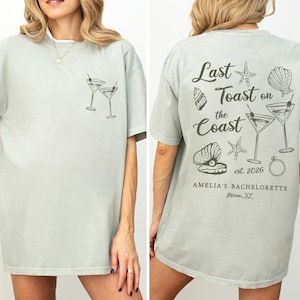 K&ouml;nnte beinhalten: Hellgr&uuml;nes T-Shirt mit Front- und R&uuml;ckendesign. Die Vorderseite zeigt zwei Martinigl&auml;ser. Auf der R&uuml;ckseite steht "Last Toast on the Coast" mit Muschel- und Seestern-Grafiken sowie "Amelia's Bachelorette Miami, FL".