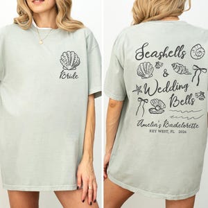 K&ouml;nnte beinhalten: Hellgr&uuml;nes T-Shirt mit einer Muschel und dem Wort "Bride" auf der Vorderseite. Auf der R&uuml;ckseite des Shirts steht "Seashells & Wedding Bells" mit Muschel- und Glockenillustrationen. Das Shirt ist f&uuml;r einen Junggesellinnenabschied in Key West, FL.