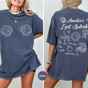 Puede incluir: Camiseta azul denim con gr&aacute;ficos de conchas blancas en la parte delantera. La parte trasera de la camiseta presenta el texto "Amelia's Last Splash" con ilustraciones de una bola de discoteca, una sirena y un anillo. Tambi&eacute;n es visible el texto "Destin, FL".