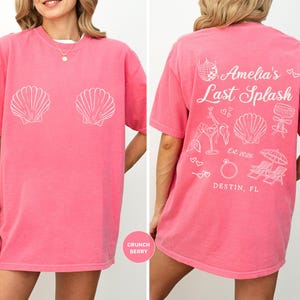 Puede incluir: Una camiseta rosa con gr&aacute;ficos de conchas blancas en la parte delantera. La parte trasera de la camiseta presenta el texto "Amelia's Last Splash" con ilustraciones tem&aacute;ticas de playa y la ubicaci&oacute;n "Destin, FL".