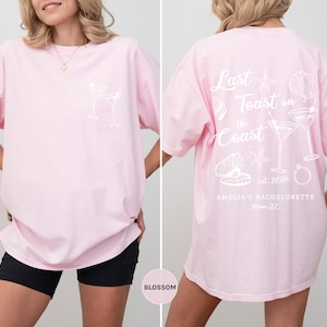 K&ouml;nnte beinhalten: Hellrosa &uuml;bergro&szlig;es T-Shirt mit wei&szlig;en Grafikdesigns. Die Vorderseite zeigt ein Cocktailglas-Design, w&auml;hrend die R&uuml;ckseite den Text "Last Toast on the Coast" mit Muschel- und Cocktail-Illustrationen zeigt. F&uuml;r Amelias Junggesellinnenabschied.