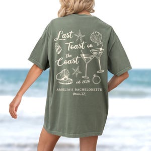 K&ouml;nnte beinhalten: Olivgr&uuml;nes T-Shirt mit wei&szlig;em Grafikdesign. Das Design enth&auml;lt den Text "Last Toast on the Coast" mit Illustrationen von Muscheln, Seesternen und Martinigl&auml;sern. Der Text "AMELIA'S BACHELORETTE Miami, FL" ist ebenfalls enthalten.