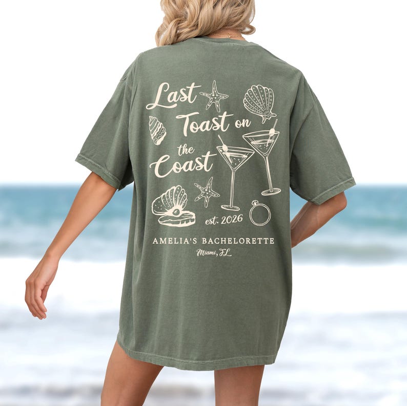 K&ouml;nnte beinhalten: Olivgr&uuml;nes T-Shirt mit wei&szlig;em Grafikdesign. Das Design enth&auml;lt den Text "Last Toast on the Coast" mit Illustrationen von Muscheln, Seesternen und Martinigl&auml;sern. Der Text "AMELIA'S BACHELORETTE Miami, FL" ist ebenfalls enthalten.