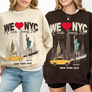 May include: Two sweatshirts, one sand-colored and one dark chocolate, each featuring a New York City design. The design includes the text "WE ❤️ NYC", a red heart, and an illustration of the city skyline with a yellow taxi. The text "Clarks Family Trip" and "NEW YORK 2025" are also present.