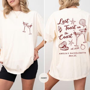 K&ouml;nnte beinhalten: Elfenbeinfarbenes T-Shirt mit Front- und R&uuml;ckendesign. Auf der Vorderseite sind zwei Martinigl&auml;ser mit Kirschen abgebildet. Auf der R&uuml;ckseite steht der Text "Last Toast on the Coast est. 2026" mit Muschel- und Seestern-Grafiken. "AMELIA'S BACHELORETTE Miami, FL" ist darunter gedruckt.