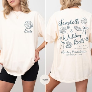 May include: Ivory-colored t-shirt with a seashell graphic and the word "Bride" on the front. The back features "Seashells & Wedding Bells" with seashell illustrations and event details. A casual, oversized fit.