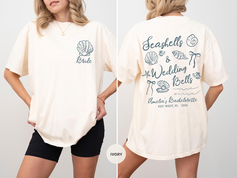 K&ouml;nnte beinhalten: Elfenbeinfarbenes T-Shirt mit einem Muschel-Motiv und dem Wort "Bride" auf der Vorderseite. Auf der R&uuml;ckseite steht "Seashells & Wedding Bells" mit Muschel-Illustrationen und Event-Details. L&auml;ssiger, &uuml;bergro&szlig;er Schnitt.