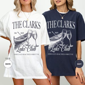 May include: White and navy blue t-shirts with a graphic design. The design includes the text "THE CLARKS Lake Club" with a boat, champagne glasses, and the text "AMELIA'S BACHELORETTE". The shirts are a relaxed fit.