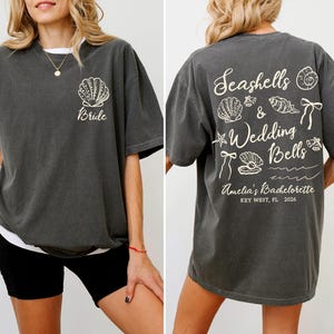 K&ouml;nnte beinhalten: Dunkelgraues T-Shirt mit einem Muschel-Motiv und dem Wort "Bride" auf der Vorderseite. Auf der R&uuml;ckseite des Shirts befinden sich Muscheln, die Worte "Seashells & Wedding Bells" und "Amelia's Bachelorette, Key West, FL, 2026".