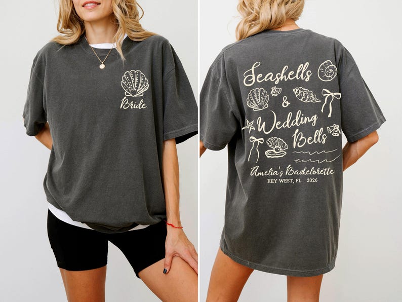 K&ouml;nnte beinhalten: Dunkelgraues T-Shirt mit einem Muschel-Motiv und dem Wort "Bride" auf der Vorderseite. Auf der R&uuml;ckseite des Shirts befinden sich Muscheln, die Worte "Seashells & Wedding Bells" und "Amelia's Bachelorette, Key West, FL, 2026".