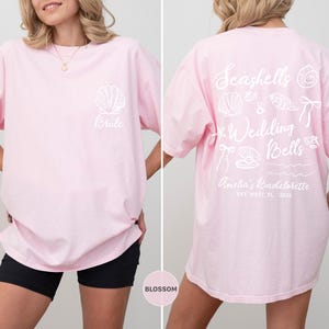 K&ouml;nnte beinhalten: Hellrosa T-Shirt mit wei&szlig;er Muschel und "Bride"-Text auf der Vorderseite. Auf der R&uuml;ckseite steht "Seashells & Wedding Bells" mit Muschel-Grafiken und "Amelia's Bachelorette, Key West, FL, 2024".
