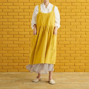 Peut inclure: Un tablier jaune moutarde avec de grandes poches et de larges bretelles. Le tablier est porté sur une blouse blanche et une jupe grise. Le fond est un mur de briques jaunes.