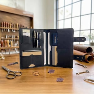 Può includere: Portfolio aperto in pelle nera con più scomparti, tra cui portapenne, fessure per carte e una tasca per passaporto. Il portfolio è aperto su una superficie di legno, mostrando la sua organizzazione interna.