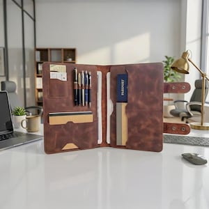 Può includere: Portfolio in pelle marrone aperto con più scomparti. Contiene penne, carte, documenti e un passaporto. Il portfolio è aperto, rivelando il suo contenuto, ed è su una superficie bianca.