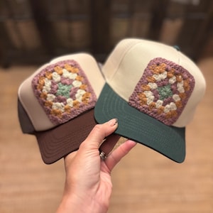 May include: Two trucker hats with a cream-colored front panel and a crocheted square design in the center. One hat has a brown brim and the other has a green brim. The hats are held up by a hand.