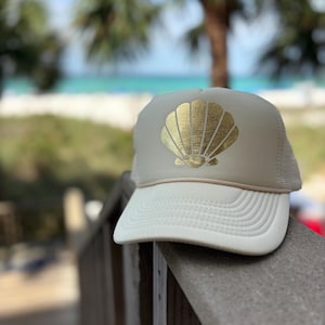 May include: A beige trucker hat with a white brim and a gold seashell design. The hat has a mesh back and a curved brim. The hat is sitting on a wooden railing with a beach and palm trees in the background.