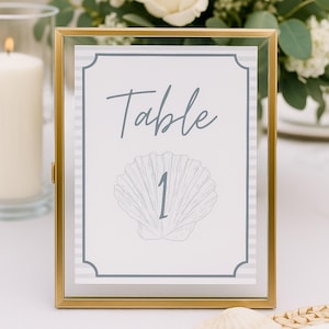 以下が含まれることがあります： 「Table」の文字が筆記体で書かれ、貝殻のイラストの下に数字の「1」が描かれた、金色のフレームのテーブルナンバーカード。 カードにはグレーと白のストライプのボーダーがあり、白いテーブルクロスの上に置かれています。