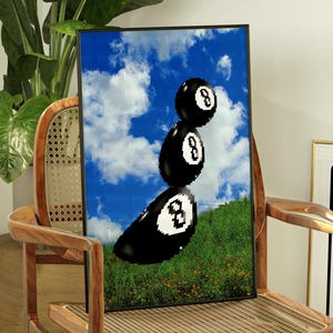 May include: A framed art print showing three black 8-ball pool balls stacked against a blue sky with clouds and green grass. The print is displayed on a wooden chair.