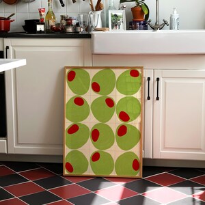 May include: A framed artwork featuring a grid of green olive shapes with red centers. The artwork is displayed in a kitchen setting with white cabinets, a farmhouse sink, and a black and red checkered floor.