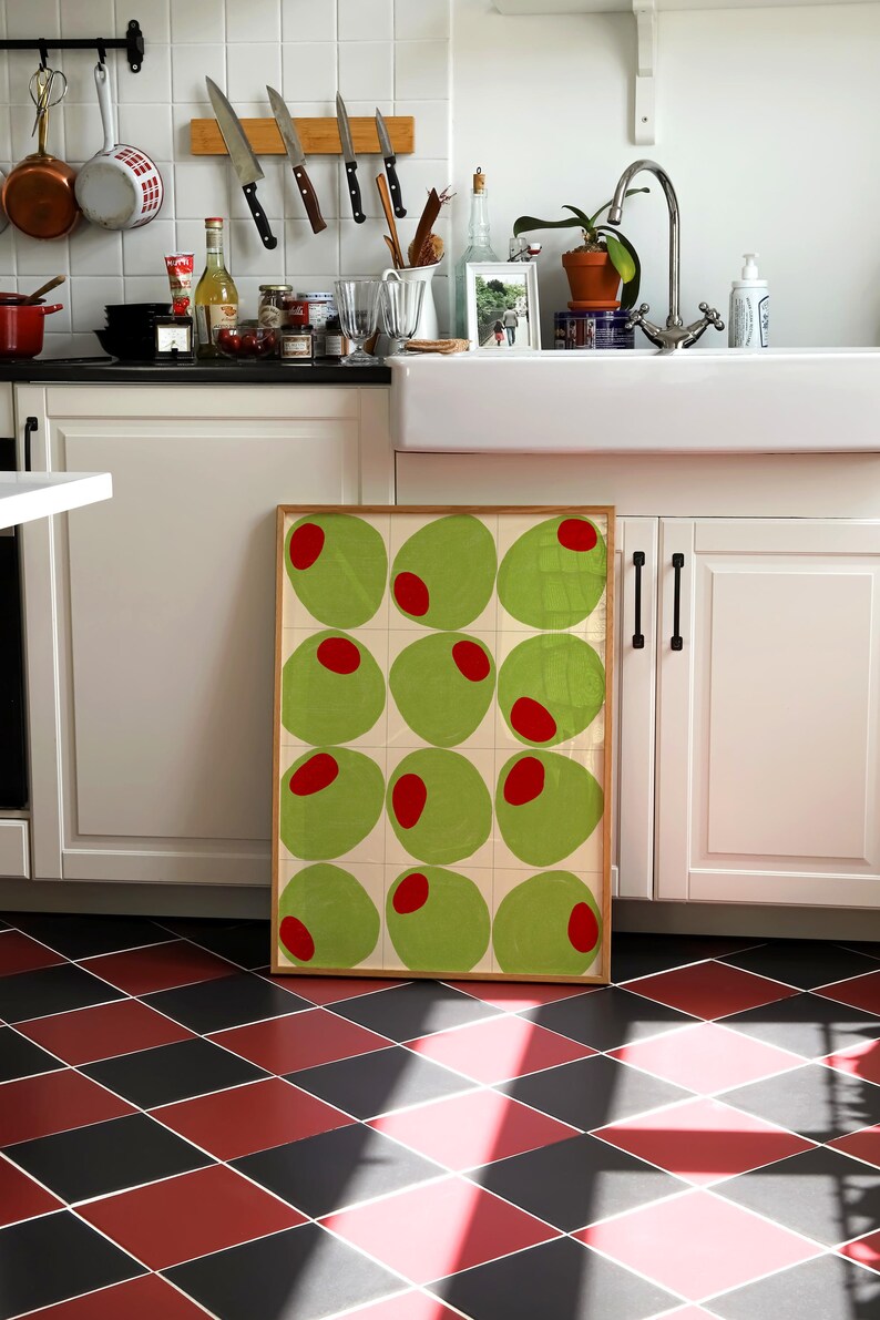 May include: A framed artwork featuring a grid of green olive shapes with red centers. The artwork is displayed in a kitchen setting with white cabinets, a farmhouse sink, and a black and red checkered floor.