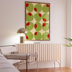 May include: A framed art print featuring a grid of green circles with red centers, resembling olives. The artwork hangs above a light pink cabinet with a ribbed design, complemented by a gold lamp and a modern chair with a gray seat.