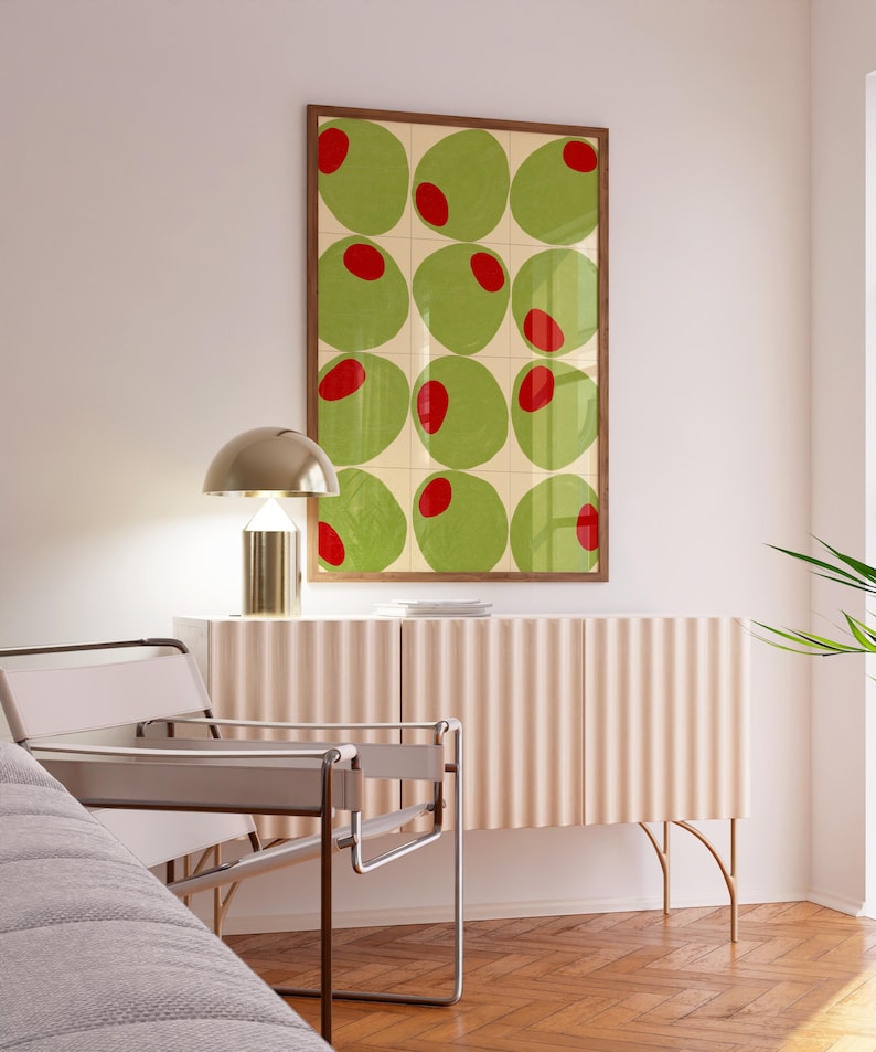May include: A framed art print featuring a grid of green circles with red centers, resembling olives. The artwork hangs above a light pink cabinet with a ribbed design, complemented by a gold lamp and a modern chair with a gray seat.