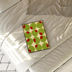 May include: A framed art print featuring a pattern of green olives with red centers on a cream background. The artwork is set against a backdrop of light gray bedding and a gray blanket.
