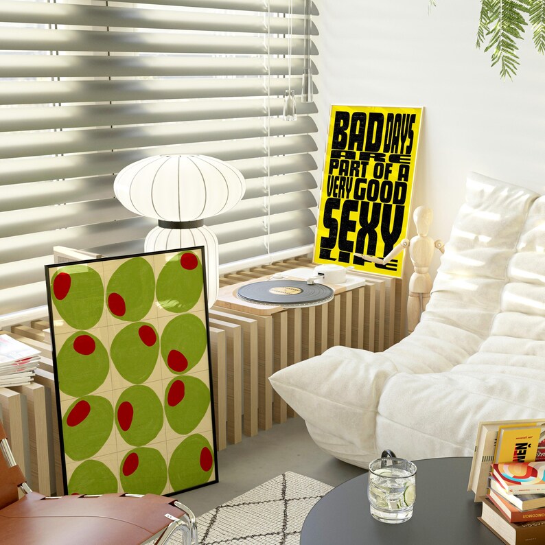 May include: A modern living room scene with a framed olive art print, a lamp, and a yellow poster that reads "BAD DAYS ARE PART OF A VERY GOOD SEXY." A record player sits on a wooden shelf. A white sofa and a black table complete the scene.