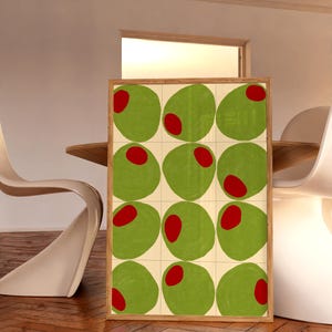 May include: A framed art print featuring a grid of twelve green olives with red centers. The olives are set against a cream-colored background, and the frame is made of light-colored wood. The artwork is displayed in a room with a wooden floor.