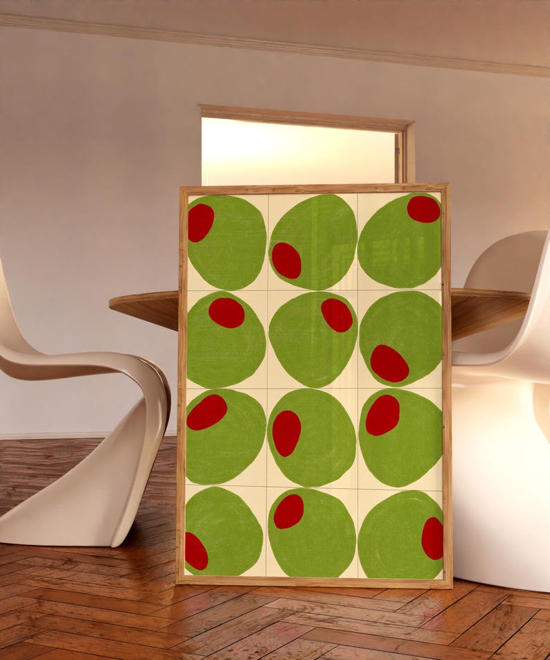 May include: A framed art print featuring a grid of twelve green olives with red centers. The olives are set against a cream-colored background, and the frame is made of light-colored wood. The artwork is displayed in a room with a wooden floor.