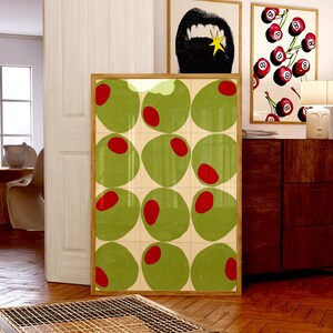 May include: A framed art print featuring a grid of green olives with red centers. The artwork is displayed in a room with other framed prints, including a black and white image of a mouth and a cherry-themed print.