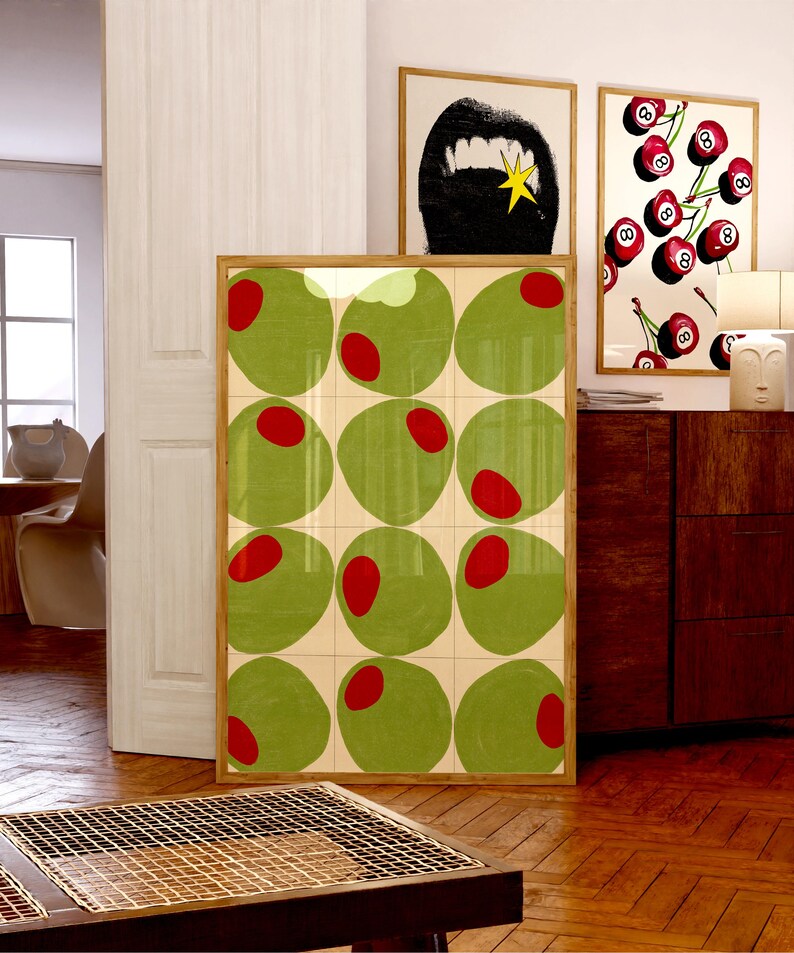 May include: A framed art print featuring a grid of green olives with red centers. The artwork is displayed in a room with other framed prints, including a black and white image of a mouth and a cherry-themed print.