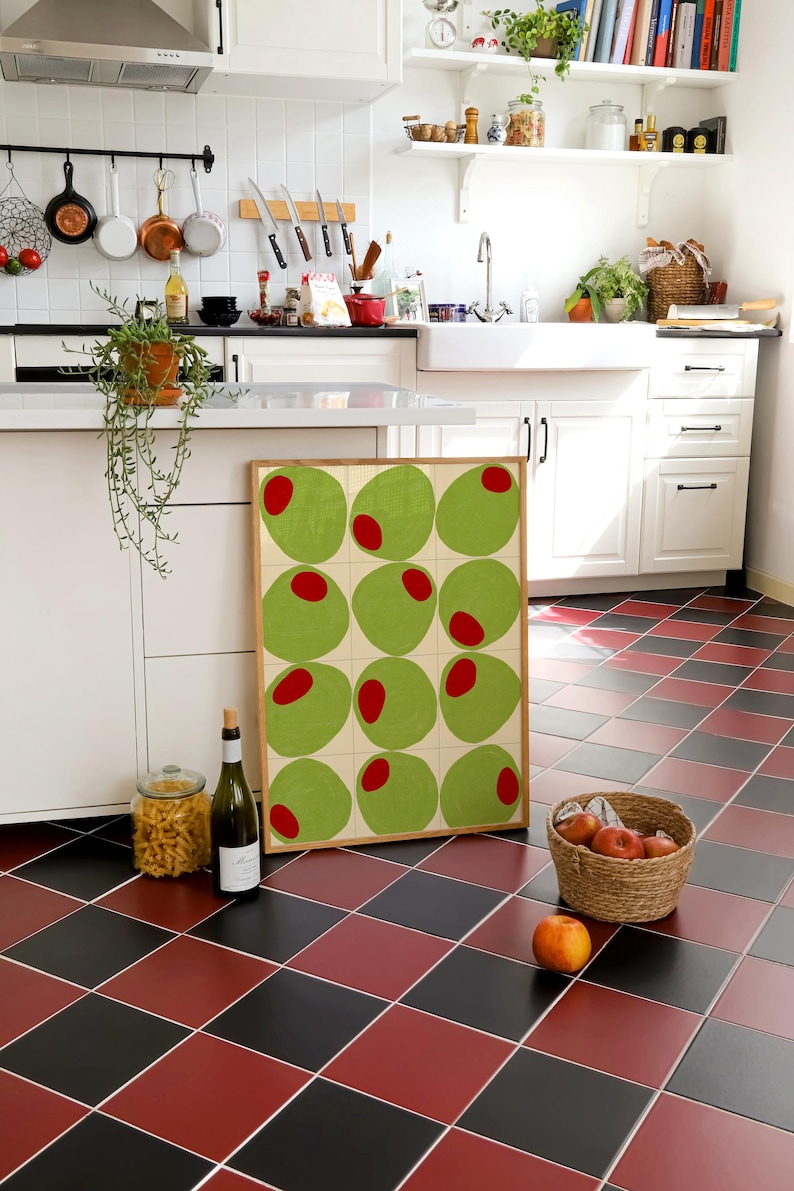 May include: A kitchen scene with a framed artwork featuring green circles with red centers. A bottle of wine, a jar of pasta, and a basket of apples sit on a red and black checkered floor. White cabinets and shelves are in the background.