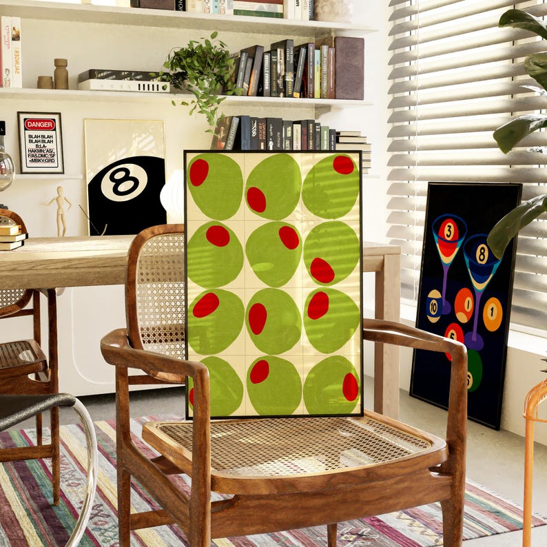 May include: A framed art print featuring a grid of green olives with red pimentos. The print is displayed on a wooden chair with a woven seat. Other art prints and decor are visible in the background.