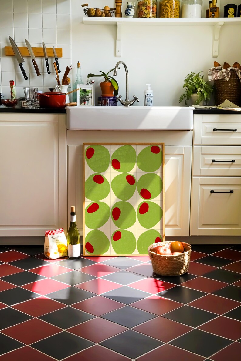 May include: A kitchen scene with a framed artwork featuring green circles with red centers. A bottle of wine, a bag of pasta, and a basket of fruit sit on the floor. The floor has a red and black diamond pattern.