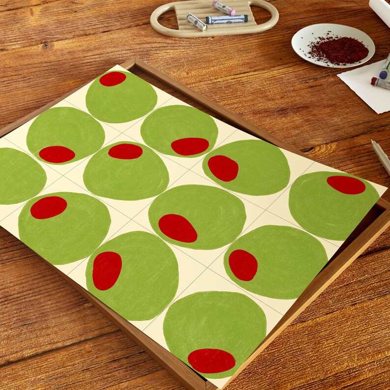 May include: A framed print featuring a pattern of green olives with red centers on a cream background. The olives are arranged in a grid pattern. The frame is a light brown color. The print is on a wooden surface.