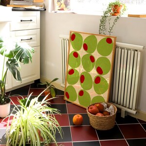 May include: A framed art print featuring a repeating pattern of green ovals with red centers. The artwork leans against a wall near a window, with a basket of peaches and potted plants on a red and black tiled floor.