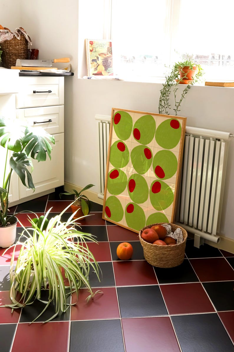May include: A framed art print featuring a repeating pattern of green ovals with red centers. The artwork leans against a wall near a window, with a basket of peaches and potted plants on a red and black tiled floor.
