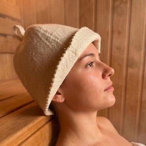 May include: A cream-colored felted wool sauna hat with a rounded crown, a small loop at the top, and a wide, folded brim. The hat features visible stitching along the seams and the edge of the brim, providing a structured shape for heat protection.