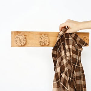 May include: A wooden coat rack with three decorative cutouts: a thermometer, a campfire, and a leaf. A brown and tan plaid shirt is hanging from the rack. The rack is made of light-colored wood.