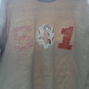 May include: A light beige sweatshirt with the number 501 sewn on in pink and red fabric. The zero is decorated with a pattern of small rainbows.
