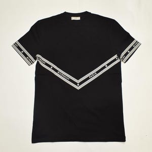 Pode incluir: T-shirt preto com design de chevron branco no peito e nas mangas. O design inclui o nome da marca "GIVENCHY" e a palavra "PARIS" em letras brancas. A camisa tem gola redonda e mangas curtas.