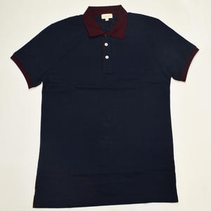 May include: A short-sleeved polo shirt in dark navy blue featuring a contrasting deep burgundy collar and sleeve cuffs. The shirt includes a two-button placket with white buttons and a small rectangular brand label located at the inner neckline.